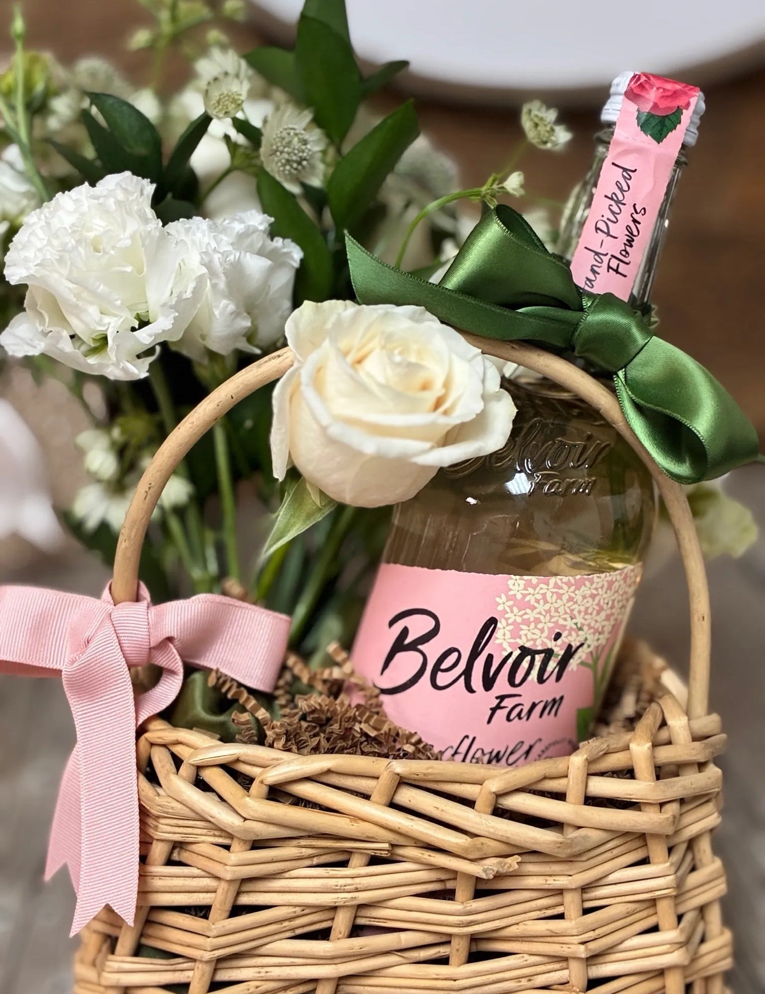 Wine & Garden Blooms Mini Basket featuring white roses, star-like astrantia, and a bottle of wine or sparkling elixir in a woven wicker basket with pink ribbon by Nifty Package Co.