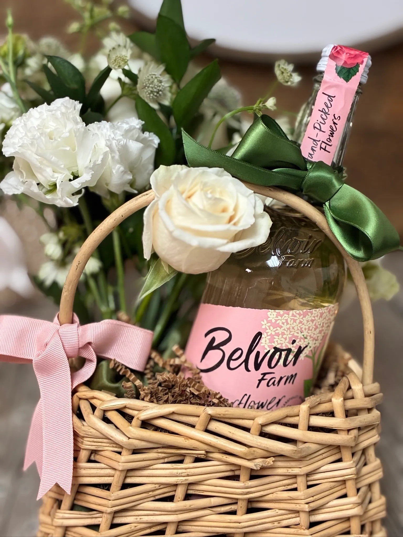 Wine & Garden Blooms Mini Basket featuring white roses, star-like astrantia, and a bottle of wine or sparkling elixir in a woven wicker basket with pink ribbon by Nifty Package Co.